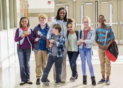 A mature African-American woman in her 40s, a teacher or parent, standing with six multi-ethnic 10 and 11 year old children in the hallway of an elementary school. The Caucasian boy in front of her wearing eyeglasses has down syndrome.